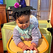 Alya a rejoint le concours — aidez-le/la à gagner de superbes lots ! toddler, child, baby, high_chair, toy, beads, witch_hat, curly_hair, indoor, wooden_furniture, playful, cute, purple_clothing, smiling, face, hands, window, curtains, bookshelf, decor