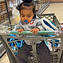 toddler, child, shopping_cart, pacifier, blanket, grocery_store, aisle, floor_tiles, clothing, hoodie, pants, socks, hand, face, hair, indoor, retail, shopping, person, seat