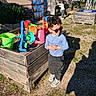child, boy, sunglasses, toys, wooden_planter, outdoor, sunlight, shadow, woman, girl, artwork, grass, gravel, building, playground, casual_clothing, nature, daytime, person, happy