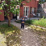 Youcef participe au concours pour gagner de l'argent avec cette photo : bike, child, costume, daytime, door, fence, flowers, grass, gravel_path, house, nature, outdoor, person, pilot_costume, play, shadow, sunglasses, sunlight, tree, window