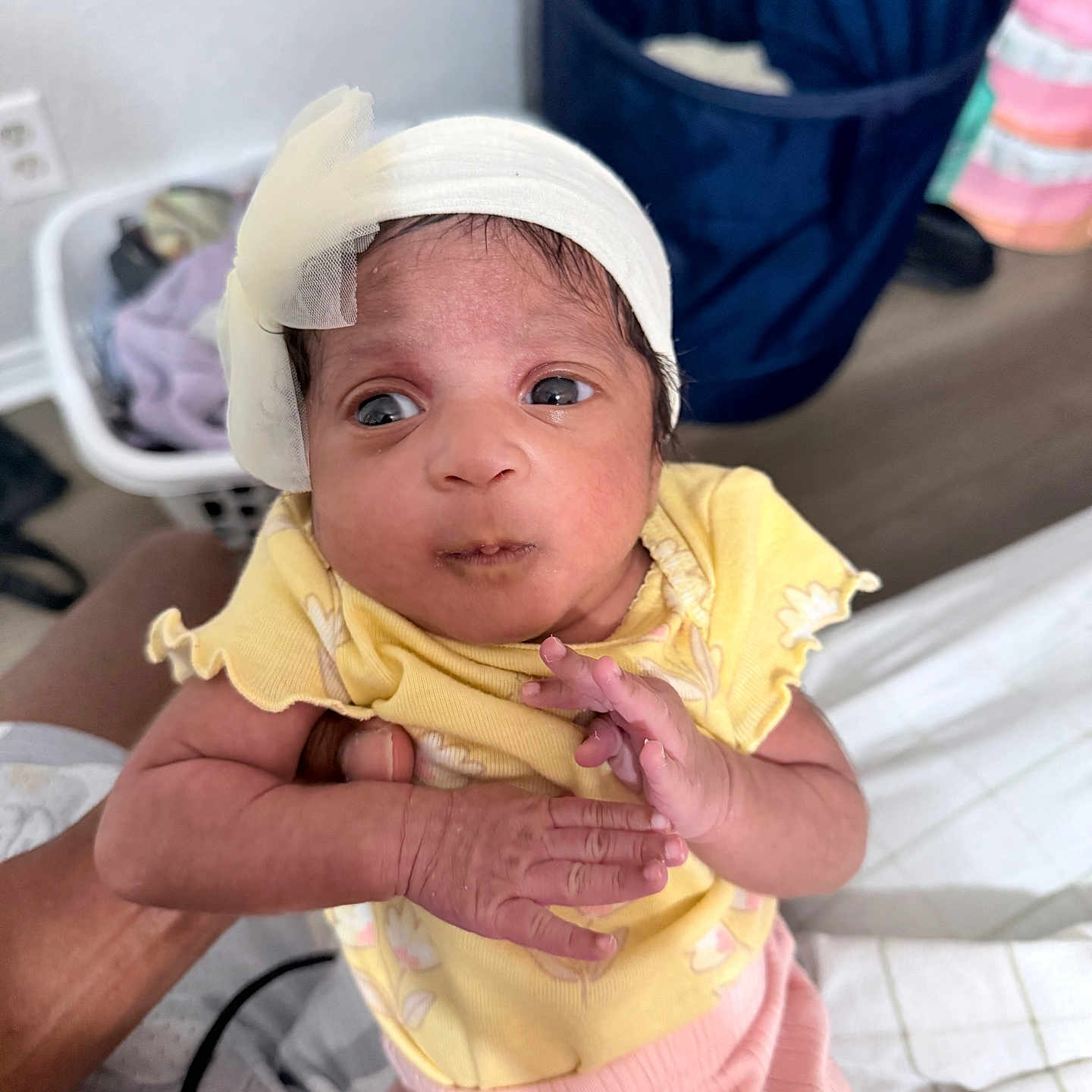 Caylani is registered to the contest to win money with this photo: baby, infant, yellow_clothing, headband, bow, hand, person, indoor, laundry_basket, fabric, pink_pants, curious_expression, skin, newborn, holding, bed, floor, home, wrinkled_skin, small_hands