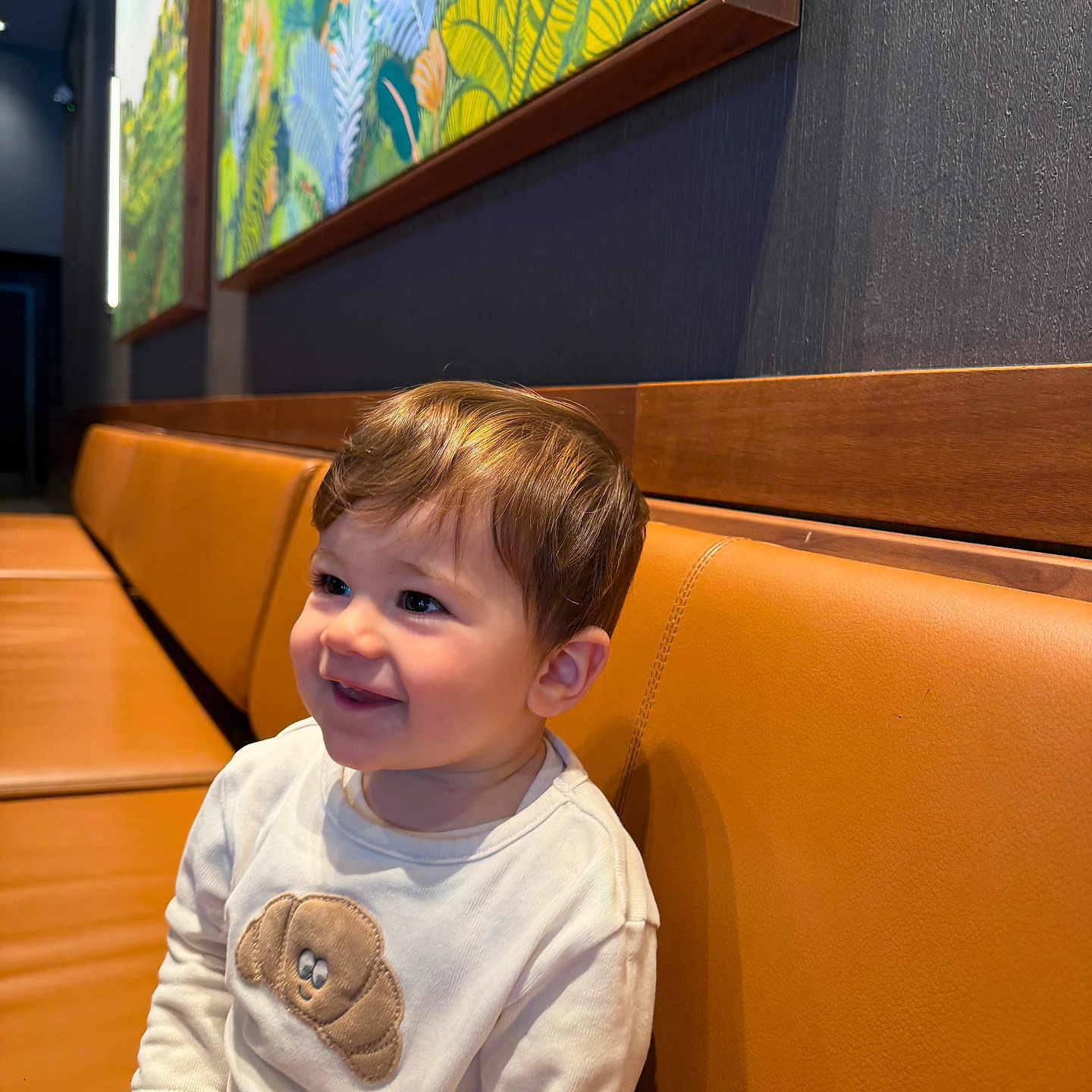 Kreg is registered to the contest to win money with this photo: toddler, child, smiling, indoor, bench, brown_hair, white_shirt, croissant_patch, orange_bench, artwork, wall_art, happy, seated, portrait, cute, cozy, casual_clothing, person, baby, furniture