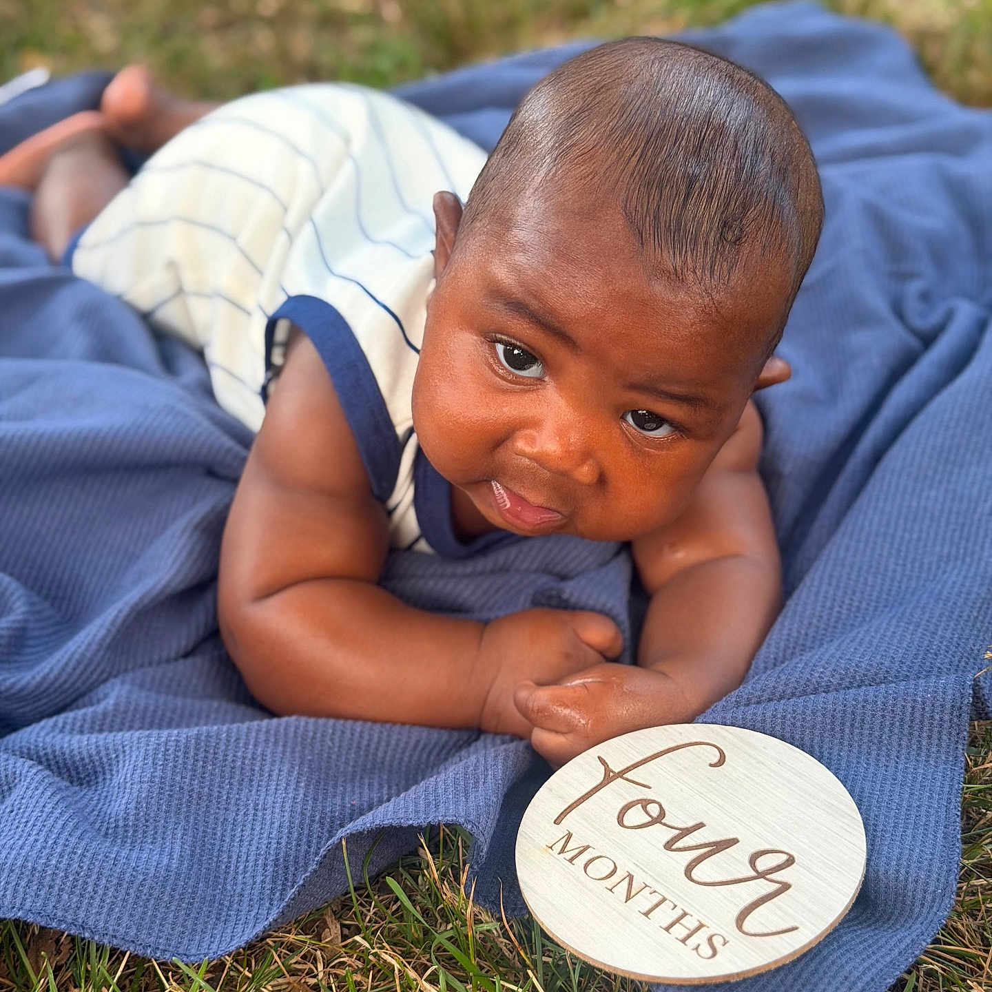 Kyaun is registered to the contest to win money with this photo: baby, infant, four_months, blue_blanket, grass, outdoor, wooden_sign, age_marker, lying_down, curious, skin, head, arms, clothing, striped_onesie, closeup, portrait, child, nature, summer