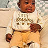 baby, child, infant, bib, yellow_pants, white_long_sleeve_shirt, animal_print, cushion, beige, soft, smiling, face, hand, footwear, sitting, indoors, portrait, cute, happy, cozy