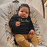 baby, child, infant, portrait, sitting, bouncer, baby_chair, smiling, wide_eyed, curly_hair, black_sweater, text_on_clothing, mustard_pants, socks, blanket, cushion, indoor, cozy, patterned_background, flawless