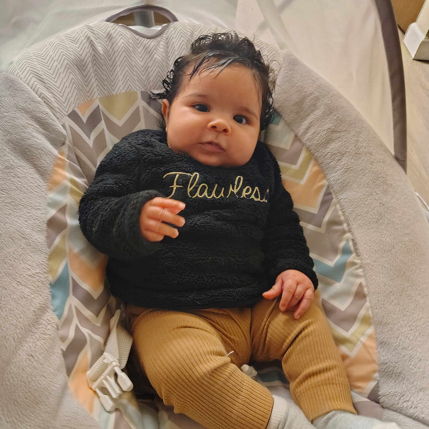 Elliyana joined the competition — help win amazing prizes! baby, child, infant, portrait, sitting, bouncer, baby_chair, smiling, wide_eyed, curly_hair, black_sweater, text_on_clothing, mustard_pants, socks, blanket, cushion, indoor, cozy, patterned_background, flawless