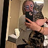 woman, cat, tabby_cat, pet, mirror_selfie, portrait, indoors, bedroom, glasses, smartphone, holding, sitting, striped_fur, green_eyes, lamp, bed, furniture, polka_dot_clothing, warm_lighting, reflection