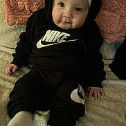 River joined the competition — help win amazing prizes! baby, child, black_clothing, nike_logo, headband, bow, sitting, blanket, cushion, indoor, soft_texture, white_socks, cute, portrait, infant, smiling, hand, face, cozy, casual
