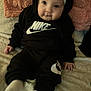 baby, child, black_clothing, nike_logo, headband, bow, sitting, blanket, cushion, indoor, soft_texture, white_socks, cute, portrait, infant, smiling, hand, face, cozy, casual
