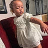 toddler, child, white_dress, curly_hair, couch, red_couch, pillow, indoor, living_room, wall_art, plush, furniture, person, portrait, exploring, cute, young_child, home, decor, balance