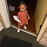 toddler, child, doorway, orange_dress, pink_shoes, hair_beads, paper, snack, indoor, floor, door, threshold, curious, standing, young_child, wood_floor, mat, casual_clothing, home, person