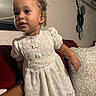 toddler, child, white_dress, lace, indoor, couch, pillow, red_couch, patterned_pillow, beaded_hair, curious_expression, person, home_decor, wall_art, hand, barefoot, portrait, young_child, furniture, casual