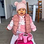 baby, child, cozy, dining_chair, floor, fuzzy_jacket, indoor, minnie_mouse, pink_hat, pink_toy, playtime, pom_pom, portrait, puffer_vest, ride_on_toy, smile, sneakers, table, toddler, wooden_chair