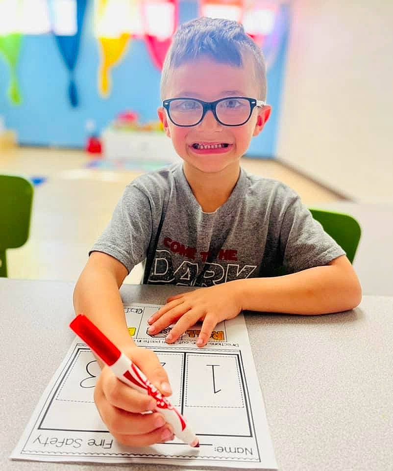 Orion is registered to the contest to win money with this photo: chair, child, design, desk, eyewear, font, fun, glasses, happy, homework, joy, nail, office_supplies, person, room, t_shirt, table, toddler, toy, writing