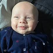 Lucas is registered to the contest to win money with this photo: baby, smile, infant, onesie, clothing, face, head, skin, lying_down, bedding, fabric, blue, cute, child, newborn, portrait, happy, soft_light, indoors, person