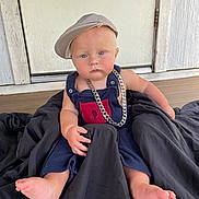 Lucas is registered to the contest to win money with this photo: baby, child, cap, chain, blue_eyes, outdoor, blanket, wooden_floor, door, overalls, serious_face, barefoot, person, portrait, sitting, clothing, accessory, fashion, young, cute