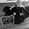 baby, child, black_and_white, chalkboard, sign, cap, overalls, floor, wooden_floor, fabric, cloth, sitting, milestone, portrait, cute, infant, person, indoors, decor, humor