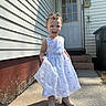 boots, child, concrete, curly_hair, daylight, door, dress, flower_pattern, happy, house, outdoor, person, side_view, sidewalk, smiling, steps, sunlight, toddler, wall, white_dress