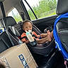 Dnero is registered to the contest to win money with this photo: toddler, car_seat, child, smiling, pacifier, bottle, car_interior, package, box, seatbelt, window, trees, road_sign, jacket, leather_seat, foot, happy, person, transportation, indoors