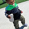 toddler, child, pacifier, shoe, jacket, green, black, pants, outdoor, concrete, grass, playground, sunny, daylight, walking, footwear, sock, curious, person, young