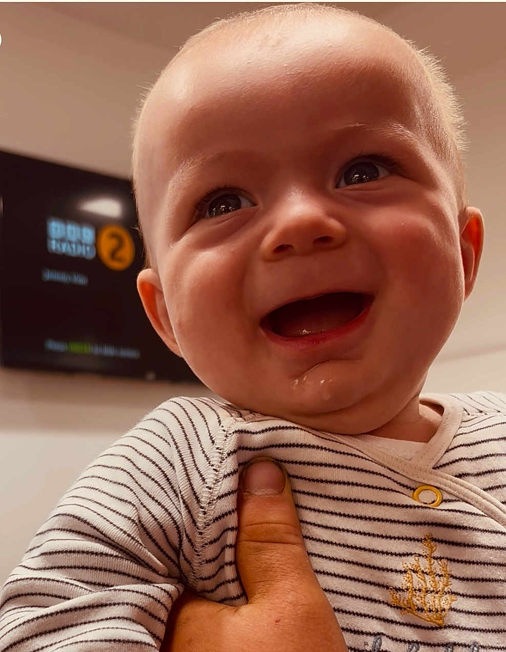 Elliott is registered to the contest to win money with this photo: baby, smiling, close_up, striped_clothing, child, happy, face, hand, indoor, portrait, infant, cute, skin, eyes, mouth, holding, background_blur, television, number, casual