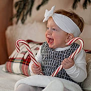 Scarlett is registered to the contest to win money with this photo: baby, candy_cane, cheerful, child, cozy, cute, decor, festive, footwear, happy, headband, holiday, indoors, person, pillow, plaid, sitting, smile, tights, white_clothing