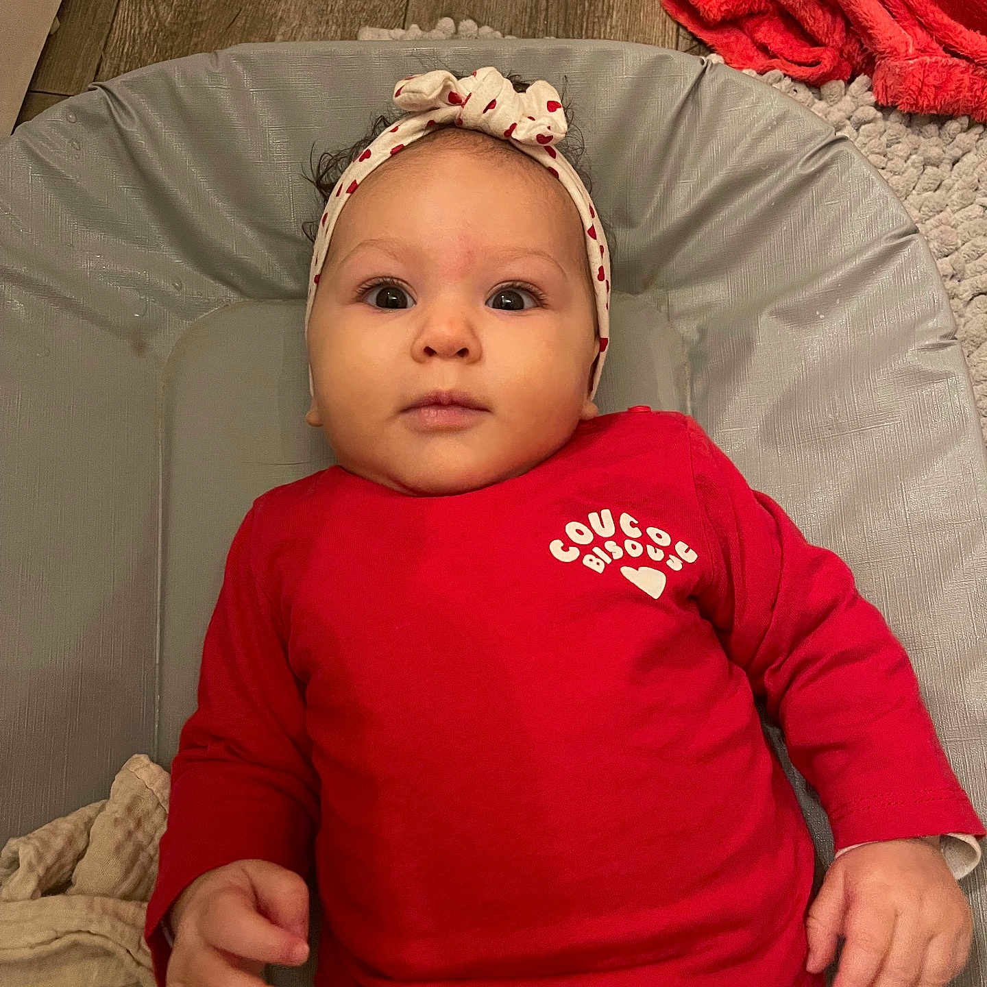Nélïa participe au concours pour gagner de l'argent avec cette photo : baby, blanket, child, clothing, curious, cushion, cute, face, floor, hand, headband, indoors, infant, lying_down, person, portrait, red_shirt, soft_texture, warm_colors, wooden_floor