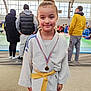 child, girl, martial_arts, yellow_belt, medal, sports, indoor, people, crowd, competition, smile, jacket, hoodie, spectators, floor, tatami, event, window, lighting, portrait