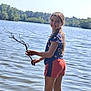child, girl, lake, water, stick, smile, outdoor, nature, trees, braid, shorts, tshirt, sunny, summer, recreation, playing, happy, standing, shore, reflection