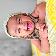 Alzaan is registered to the contest to win money with this photo: baby, smiling, knitted_hat, quilt, yellow, bedroom, happy, child, infant, cute, face, skin, arm, hand, lying_down, soft_focus, button, textile, headwear, indoors
