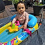 casual_clothing, child, curly_hair, daylight, deck, expression, flower_toy, garden, hose, inflatable_pool, outdoor, person, playtime, potted_plants, residential_area, summer, sunny, toddler, toy, water