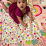 baby, child, purple_dress, play_mat, rainbows, polka_dots, colorful, infant, cute, smiling, indoors, floor, toddler, patterned, clothing, person, happy, portrait, lying_down, young_child