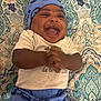 baby, infant, laughing, smiling, blue_hat, blue_pants, clothing, patterned_blanket, happy, joyful, cute, lying_down, hands_clasped, portrait, child, indoors, fabric, text_on_clothing, close_up, human