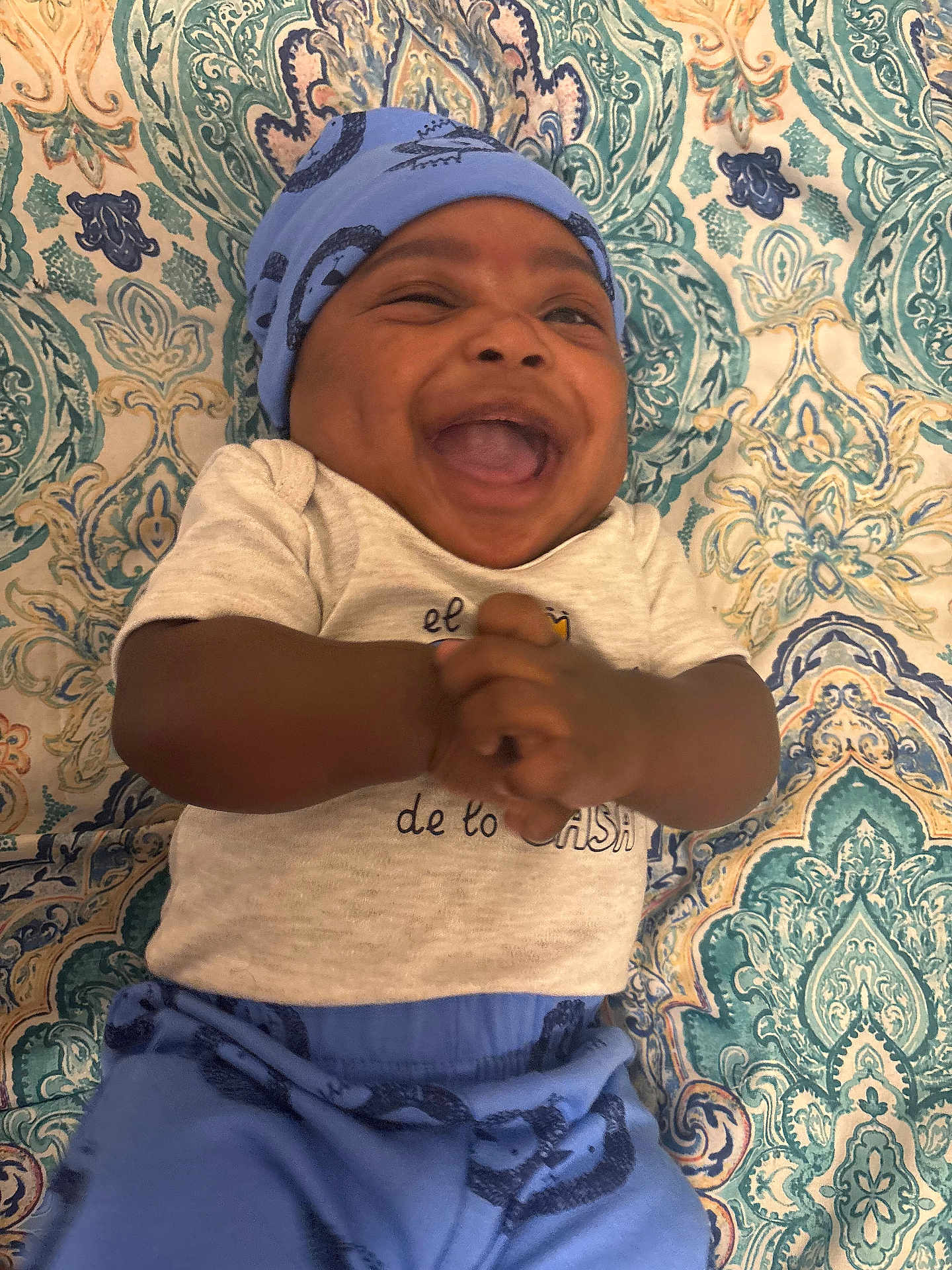 Khazir joined the competition — help win amazing prizes! baby, infant, laughing, smiling, blue_hat, blue_pants, clothing, patterned_blanket, happy, joyful, cute, lying_down, hands_clasped, portrait, child, indoors, fabric, text_on_clothing, close_up, human