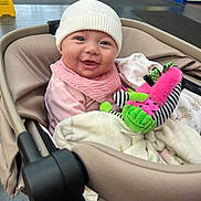 Annie joined the competition — help win amazing prizes! baby, smiling, stroller, indoor, hat, plush_toy, child, blanket, pink_bib, happy, person, public_space, floor, caution_sign, clothing, seated, face, headwear, toy, infant