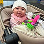 baby, smiling, stroller, indoor, hat, plush_toy, child, blanket, pink_bib, happy, person, public_space, floor, caution_sign, clothing, seated, face, headwear, toy, infant