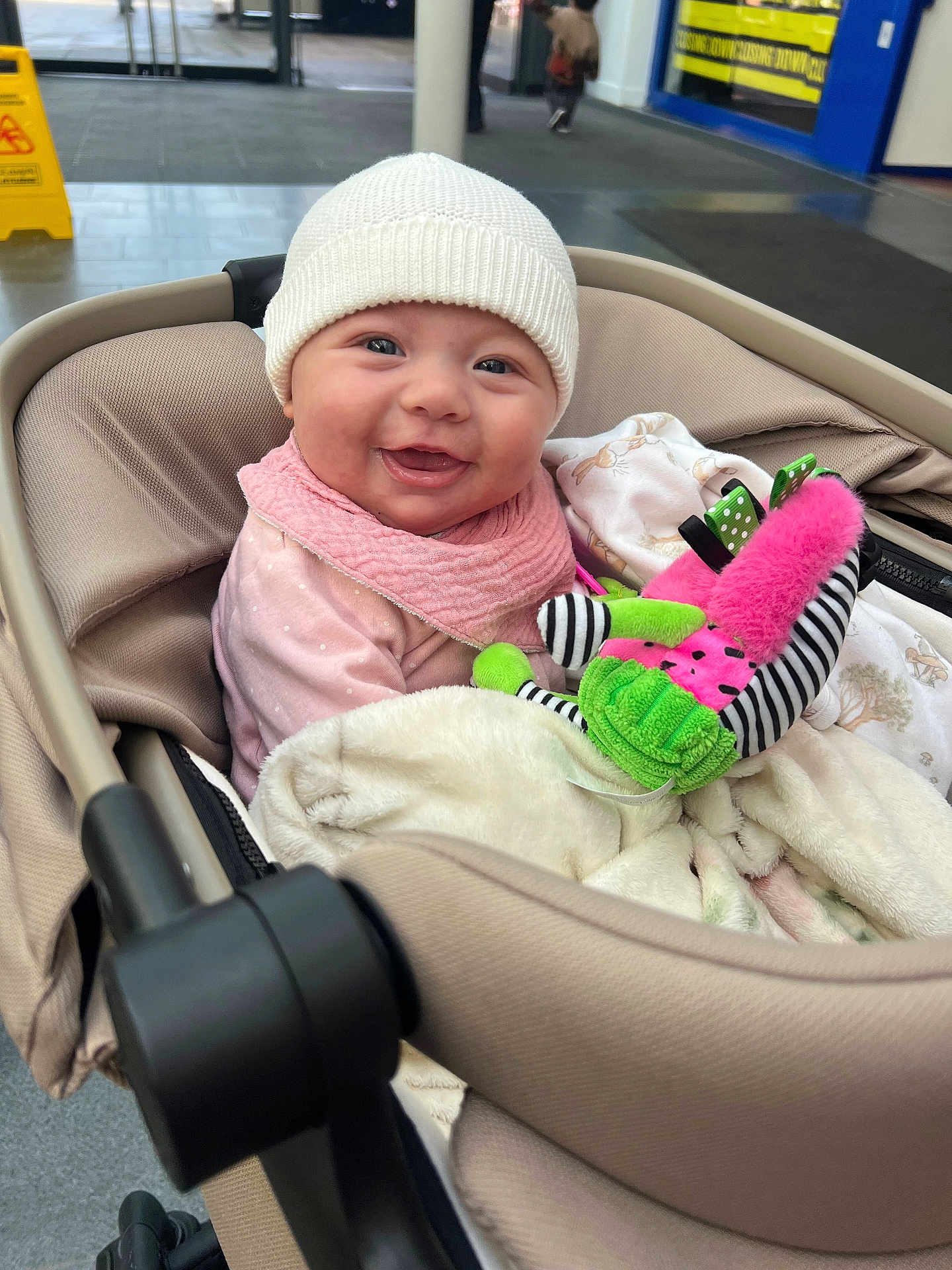 Annie joined the competition — help win amazing prizes! baby, smiling, stroller, indoor, hat, plush_toy, child, blanket, pink_bib, happy, person, public_space, floor, caution_sign, clothing, seated, face, headwear, toy, infant