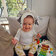 Eliott participe au concours pour gagner de l'argent avec cette photo : baby, child, smiling, hat, stuffed_animal, toy, musical_keyboard, cushion, sofa, indoor, window, sunlight, clothing, pants, foot, hand, happy, infant, playful, home