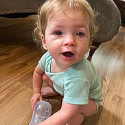 Scarlette is registered to the contest to win money with this photo: toddler, child, baby_bottle, floor, wood_floor, onesie, green_clothing, indoor, sofa, furniture, curly_hair, barefoot, person, crouching, holding, face, mouth_open, blue_eyes, skin, home