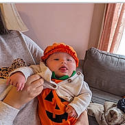 Alana is registered to the contest to win money with this photo: baby, infant, pumpkin_costume, orange_clothing, hat, tongue_out, holding, person, sweatshirt, leopard_print, couch, blanket, living_room, curtains, window, hand, face, cute, indoor, cozy