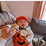 baby, infant, pumpkin_costume, orange_clothing, hat, tongue_out, holding, person, sweatshirt, leopard_print, couch, blanket, living_room, curtains, window, hand, face, cute, indoor, cozy