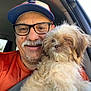 man, dog, glasses, hat, car, seatbelt, orange_shirt, smiling, portrait, pet, fluffy_dog, window, interior, companion, casual, beard, mustache, daylight, close_up, happy