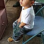 child, boy, sitting, chair, indoor, curious, white_shirt, camouflage_pants, tan_boots, adult, green_shoes, floor, concrete, folding_chair, table, basket, phone, can, casual, portrait