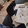 kitten, black_cat, couch, books, papers, remote_control, blanket, indoor, tile_floor, curious, stack, reading_material, cozy, pet, feline, home, furniture, small_cat, sitting, domestic