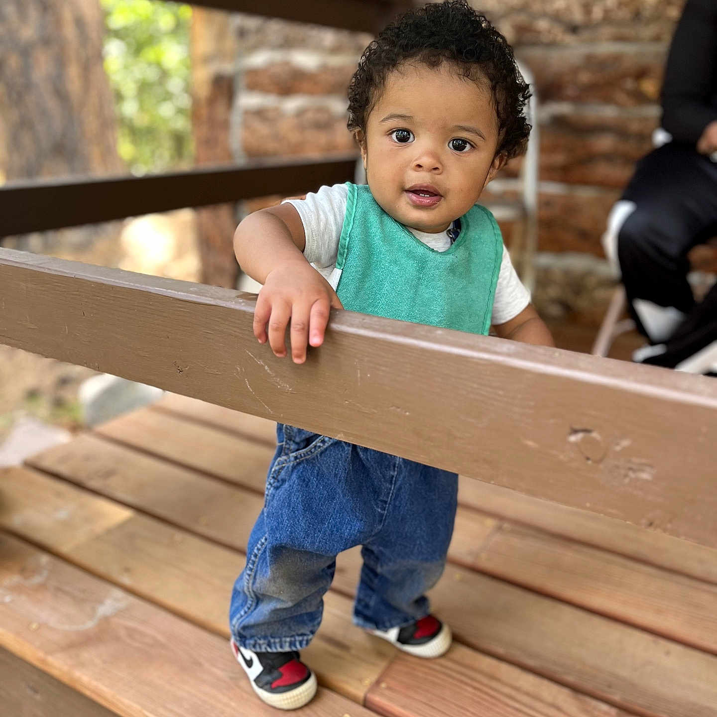 Justice joined the competition — help win amazing prizes! toddler, child, curly_hair, blue_jeans, sneakers, teal_bib, wooden_deck, railing, outdoor, person, curious_expression, hand, fence, portrait, daylight, casual_clothing, nature_background, blurred_background, standing, baby