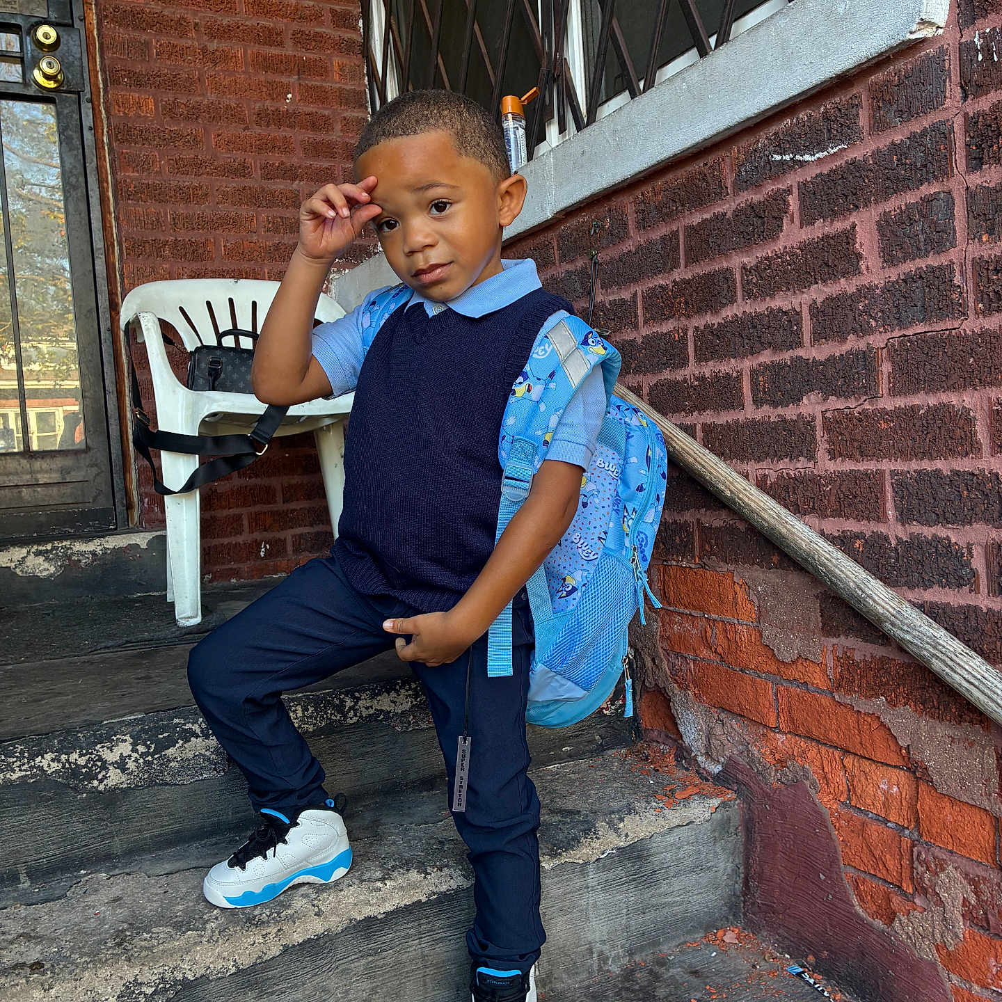 Trevon joined the competition — help win amazing prizes! child, boy, backpack, steps, brick_wall, plastic_chair, door, window, sidewalk, outdoor, casual_clothing, sneakers, pose, young, person, urban, curious, confident, daylight, playful