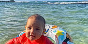 Zaizen is registered to the contest to win money with this photo: baby, child, inflatable_ring, ocean, water, tattoos, bracelets, sky, clouds, waves, red_shirt, swimming, summer, beach, sunlight, outdoor, cute, holding, person, fun