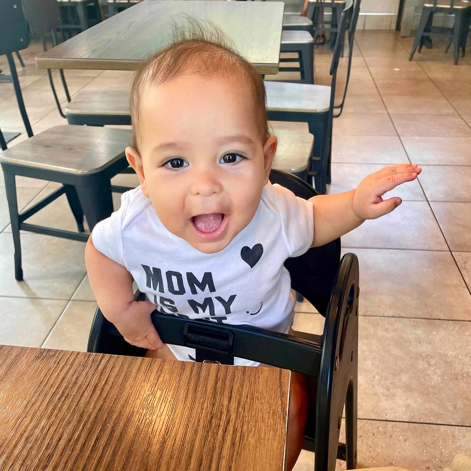 Zaizen is registered to the contest to win money with this photo: arm, baby, chair, child, clothing, cute, eyes, furniture, hair, happy, high_chair, indoor, infant, mouth_open, person, restaurant, smiling, table, tile_floor, white_shirt