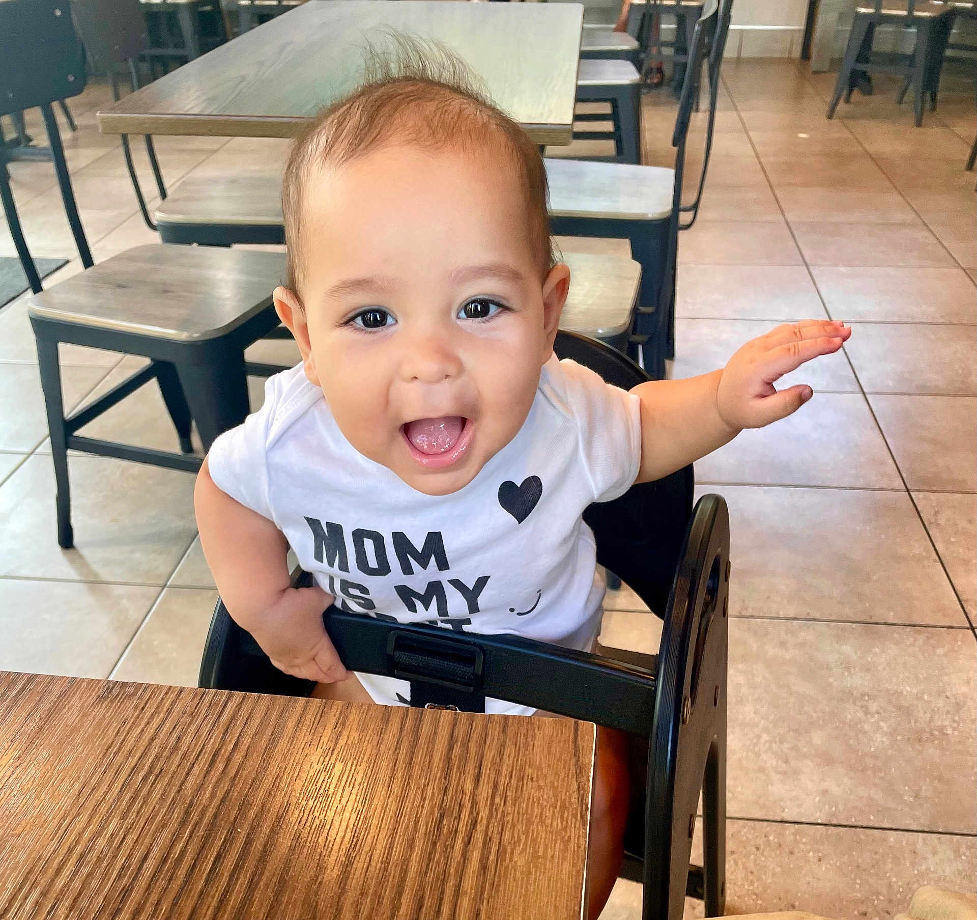 Zaizen is registered to the contest to win money with this photo: baby, child, high_chair, white_shirt, smiling, happy, indoor, restaurant, table, chair, tile_floor, cute, person, infant, arm, mouth_open, eyes, hair, clothing, furniture