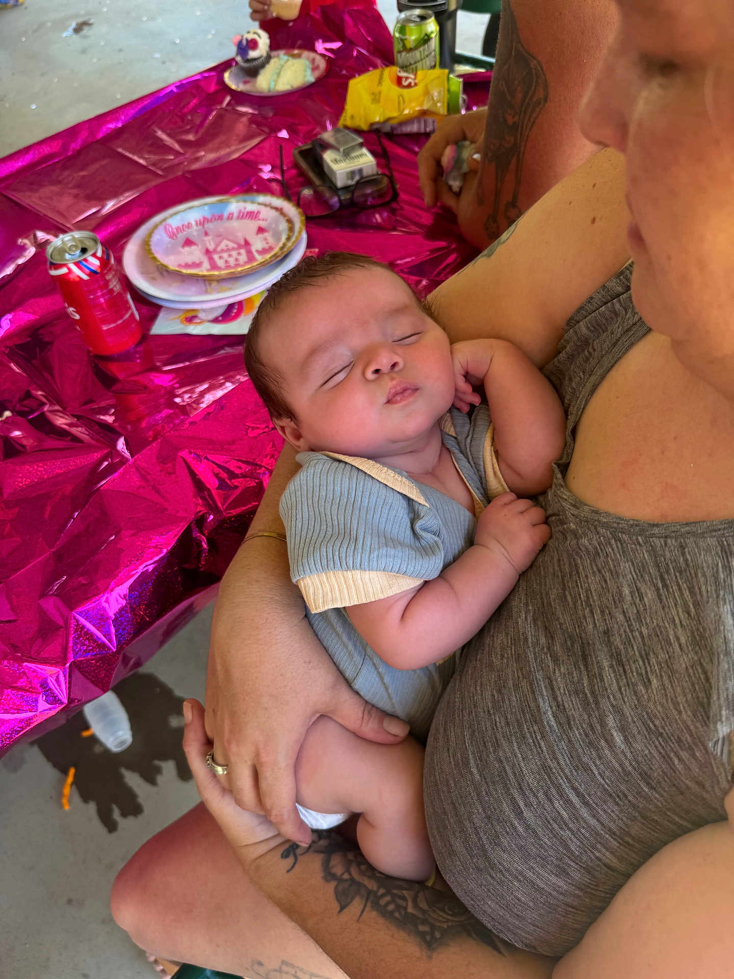Taylyn is registered to the contest to win money with this photo: baby, sleeping, infant, person, adult, tattoo, arm, table, party, snacks, soda_can, plate, napkin, clothing, hand, celebration, pink_wrapping, food, indoor, casual